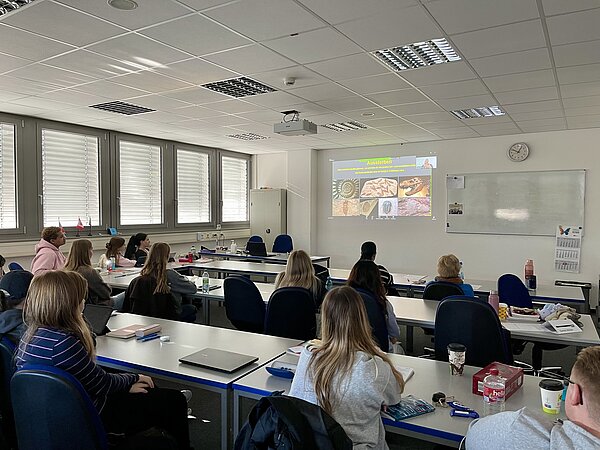 Euro Akademie Aschaffenburg will Klimaschule werden Studierende sitzen in einem modernen Klassenraum der Euro Akademie und verfolgen aufmerksam eine Präsentation, die an die Wand projiziert wird. Auf der Folie sind Bilder von Fossilien und der Begriff „Aussterben“ zu sehen.