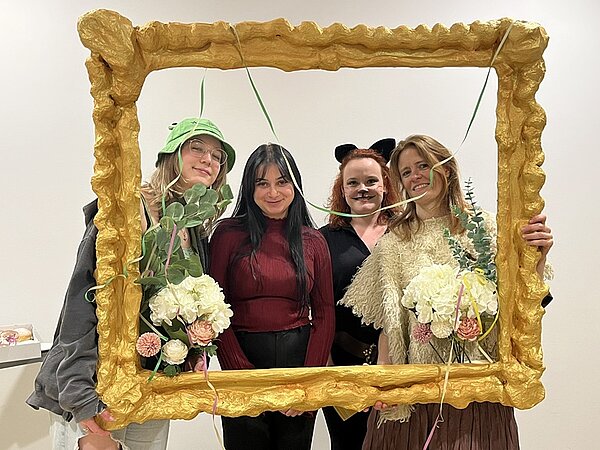 Vier weibliche Studierende schauen lächelnd in ihren Faschingskostümen durch einen übergroßen selbst gestalteten Bilderrahmen und posieren für ein Foto.