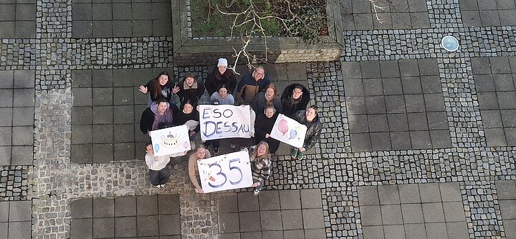 Gruppe von Schüler*innen steht auf einem gepflasterten Innenhof und hält selbst gestaltete Plakate mit „ESO Dessau“ und „35“ in die Kamera.