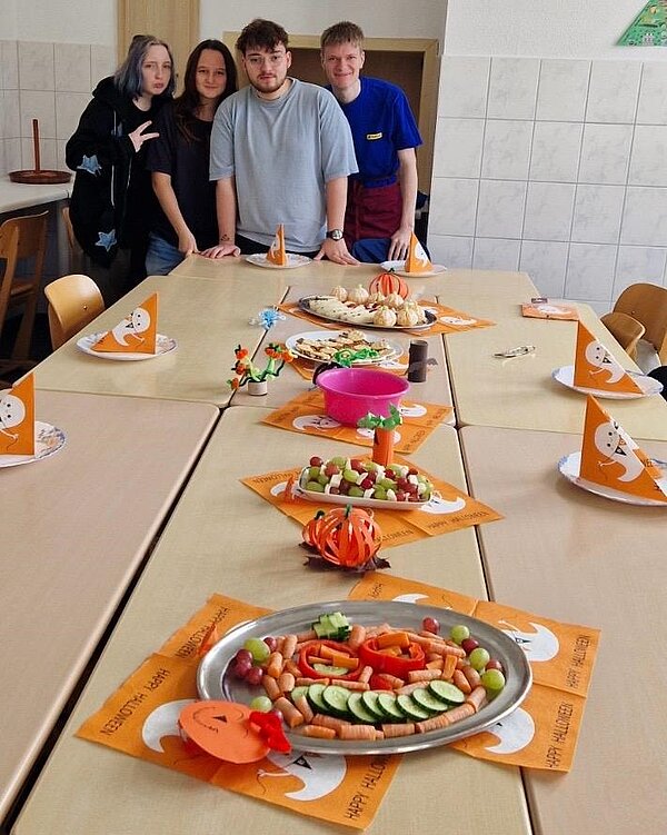 Gesund und gruselig: Kitaessen zu Halloween Die Auszubildenden stehen an ihrer gedeckten Halloween-Tafel, mit ihren zubereiteten Snacks und posieren gut gelaunt für ein Foto.