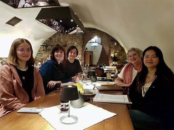 Alumnitreffen 2025 Ehemalige Auszubildende der Euro Akademie Aschaffenburg sitzen an einem Tisch im Restaurant und posieren, gemeinsam mit einer Lehrerin, lächelnd für ein Gruppenfoto. Vor ihnen liegen Speisekarten, Gläser mit verschiedenen Getränken, Besteck, eine gelbe Blume in einer kleinen Vase und eine kleine Tischlampe.