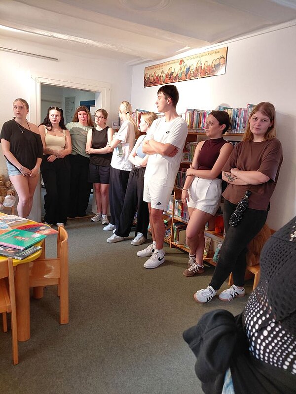 Eine Gruppe von Jugendlichen steht eng nebeneinander in einem hellen Raum in dem ein niedriger Tisch mit Kinderstühlen steht. Hinter der Gruppe stehen Regale mit Büchern.