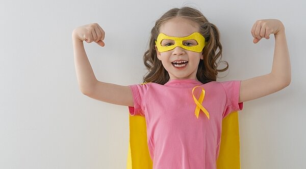 Ein kleines Mädchen in einem rosa T-Shirt und mit einem Heldenumhang und einer entsprechenden Maske über den Augen zeigt uns ihre Muskeln. An ihrem T-Shirt prangt die gelbe Schleife, die für Solidarität mit Krebs bei Kindern steht.