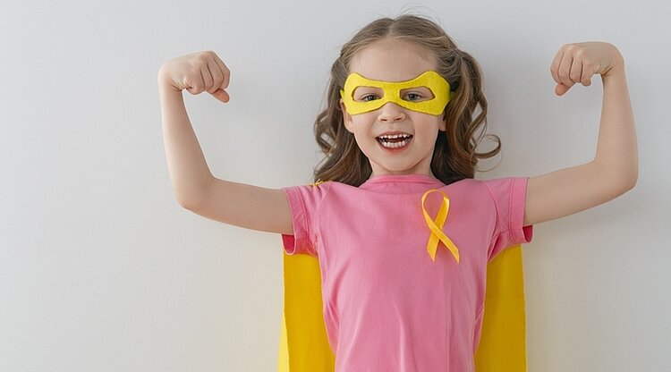 Ein kleines Mädchen in einem rosa T-Shirt und mit einem Heldenumhang und einer entsprechenden Maske über den Augen zeigt uns ihre Muskeln. An ihrem T-Shirt prangt die gelbe Schleife, die für Solidarität mit Krebs bei Kindern steht.