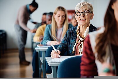 In einem Klassenraum sitzen jüngere und ältere Menschen. In der Mitte sitzt eine ältere Frau mit kurzem grauen Haaren und einem bunten Schal umgebunden, die einen Stift in der Hand hält und etwas schreibt.