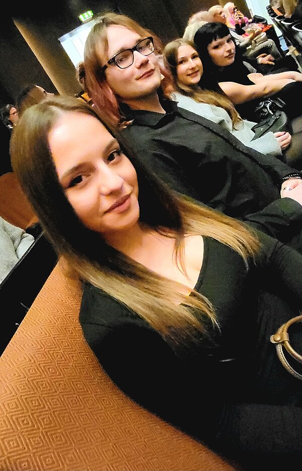 Mehrere Schülerinnen und Schüler sitzen im Opernhaus Hannover, auf gepolsterten Sitzen mit einem orangefarbenen, gemusterten Stoffbezug. Die vorderste Person trägt ein schwarzes Oberteil und hat lange, glatte, hellbraune Haare. Im Hintergrund sind weitere Zuschauer zu sehen, die ebenfalls auf den Sitzreihen nebeneinander sitzen. Die Beleuchtung ist gedämpft, typisch für eine Theaterumgebung vor Beginn oder während einer Vorstellung. Die Szene zeigt den Besuch von Schülerinnen und Schülern der Euro Akademie Hannover bei der Ballettaufführung „Goldberg“.