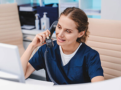 Eine Frau in medizinischer Kleidung sitzt an einem Schreibtisch und telefoniert mit einem Headset. Sie trägt eine dunkelblaue Uniform und konzentriert sich auf ihren Computerbildschirm. Im Hintergrund sind medizinische Utensilien und Flaschen zu sehen.