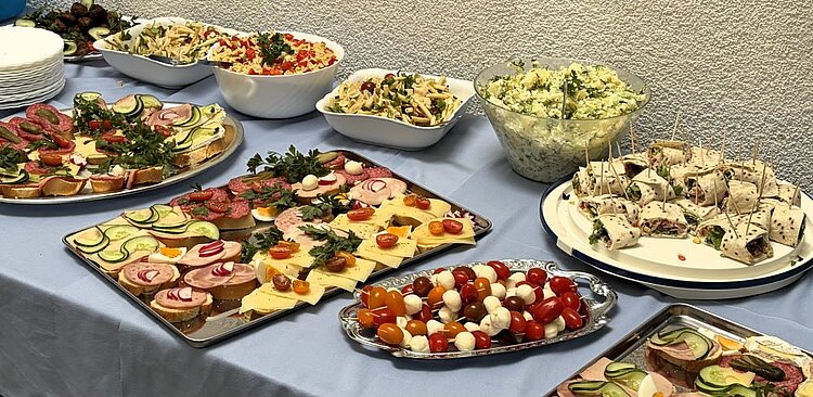 Ein üppig gedeckter Buffettisch mit einer Vielzahl an kalten und warmen Speisen. Dazu gehören verschiedene Salate, Aufschnittplatten mit Wurst, Käse und Gemüse, Antipasti, gefüllte Teigtaschen sowie Dips und Beilagen. Alles ist ansprechend auf Platten und in Schüsseln angerichtet.