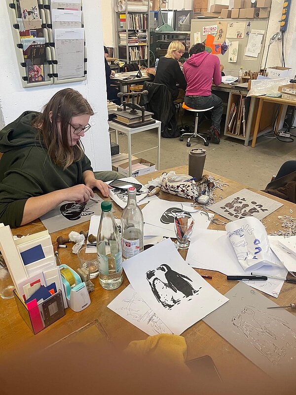 In einem Atelier sitzt eine Person mit langen braunen Haaren und Brille an einem großen Holztisch und arbeitet konzentriert an einer Zeichnung. Auf dem Tisch liegen Werkzeuge, Papierbögen mit Zeichnungen – darunter Pinguine – sowie Flaschen und Gläser. Im Hintergrund arbeiten zwei weitere Personen an ihren Tischen, während Regale mit Materialien und Wände mit Zeichnungen und Notizen den Raum prägen.