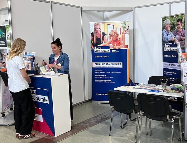 Messestand mit mehreren Roll-up-Bannern und einem weißen Tresen mit blauem Aufdruck. Zwei Frauen stehen am Tresen, eine davon hinter dem Tresen, die andere davor. Auf den Bannern sind Fotos von Menschen in Lernsituationen sowie der Schriftzug 'Euro Akademie' und weitere Informationen zu sehen. Auf dem Tisch rechts liegen Prospekte und Stifte. Im Hintergrund stehen schwarze Stühle um einen Tisch.