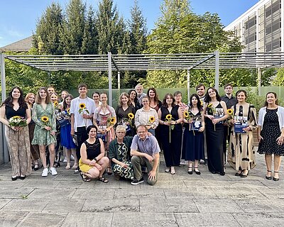 Eine Gruppe von jüngeren und älteren Menschen steht und kniet auf einer Steinterrasse. Sie sind festlich gekleidet und einige haben neben einer blauen Zeugnismappe auch Sonnenblumen in den Händen.