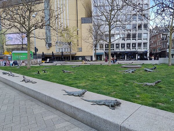 Ein öffentlicher Platz in der Stadt mit einer Installation aus kleinen Skulpturen, die an Kaimane erinnern. Im Hintergrund sind Gebäude und Bäume zu sehen.