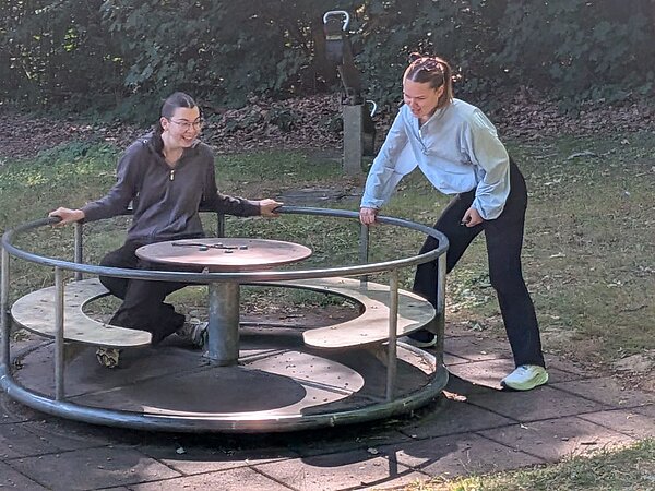 Zwei Frauen auf einem drehbaren Karussell auf einem Spielplatz im Freien. Eine Frau sitzt auf dem Karussell, während die andere es in Bewegung hält. Im Hintergrund sind Bäume und eine Wippe zu sehen.