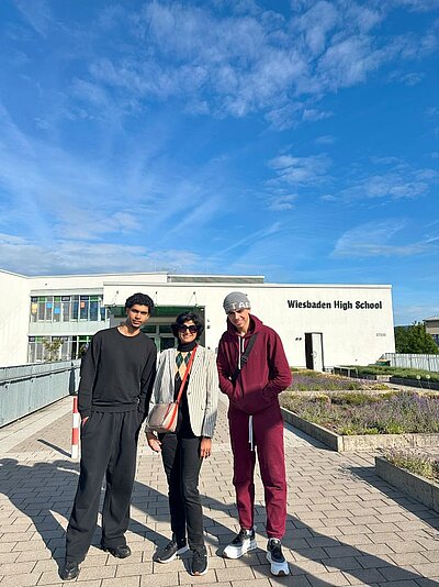 Drei Personen stehen vor dem Eingang der Wiesbadener High School. Das Gebäude ist modern und hell, umgeben von gepflegten Beeten mit Lavendelpflanzen. Der Himmel ist klar und blau.