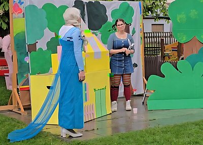 Zwei Personen stehen auf einer Rasenfläche vor bemalten Kulissen, die Bäume und ein gelbes Haus darstellen. Die linke Person trägt ein langes, hellblaues Kleid mit Schleier und Umhang, während die rechte Person ein kurzes, blaues Kleid mit weißen Knöpfen, gestreiften Strumpfhosen und weißen Schuhen trägt. Im Vordergrund steht ein weißer Becher auf dem Boden. Die Szene wirkt wie eine Theateraufführung im Freien.