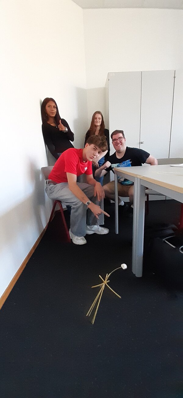 „Die Neuen“ sind da – Euro Akademie Mainz begrüßt neue Klasse der Fremdsprachenkorrespondenten Eine Gruppe von vier Personen sitzt am Tisch, im Vordergrund ist ein gescheiterter Spaghetti-Turm mit Marshmallow zu sehen, über den alle lachen.