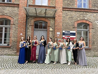 Eine Gruppe von elf Frauen steht vor einem Gebäude der Euro Akademie. Zehn von ihnen tragen festliche Kleider und halten Blumen sowie Urkunden in den Händen – offenbar frisch absolvierte Absolventinnen. Links steht eine Frau in schwarzer Kleidung mit roter Jacke, vermutlich eine Lehrkraft. Alle lächeln in die Kamera.