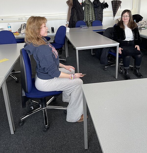 Zwei weibliche Lehrkräfte sitzen in einem büroähnlichen Raum mit mehreren grauen Schreibtischen und blauen Stühlen. Die Person links sitzt im Profil, trägt einen blauen Kapuzenpullover und eine graue Jogginghose und hält ein Handy in der Hand. Die Person rechts blickt in die Kamera und trägt eine schwarze Jacke und einen schwarzen Rock. Im Hintergrund hängen Mäntel und ein Regenschirm an der Wand. Der Boden ist mit Teppich ausgelegt, und auf den Schreibtischen stehen verschiedene Gegenstände wie Wasserflaschen und Bücher.