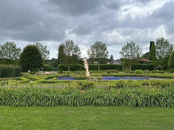 Unterwegs mit Wanderschuhen und einem Erkundungsbuch Gepflegter Park mit Hecken, Blumen und einem auffälligen Kunstobjekt in der Mitte, das wie eine schräg gestapelte Säule aussieht. Im Hintergrund stehen Bäume und Häuser.