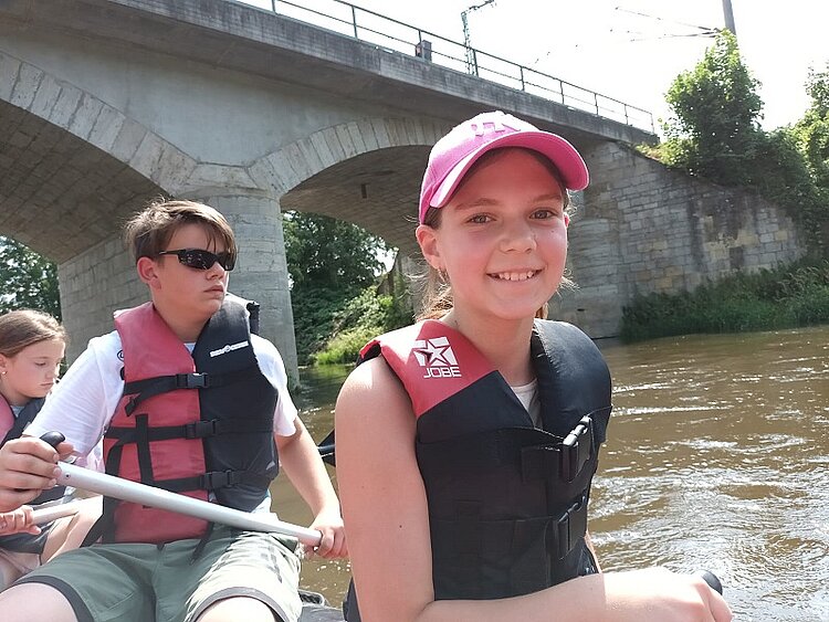 MDR berichtet über Sprachferien Ein Mädchen mit Baseballkappe und Schwimmweste grinst in die Kamera. Sie sitzt auf einem Boot das gerade auf einem Fluss eine Brücke unterquert. Hinter ihr sitzt ein Junge mit Sonnenbrille, dahinter noch ein Mädchen. Alle drei Jugendlichen haben ein Ruder in der Hand und auch die anderen beiden tragen Schwimmwesten.