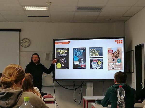 MeKoSax-Projekt „Trickfilmproduktion“ Eine Frau präsentiert verschiedene Plakate auf einem Bildschirm und davor sitzen mehrere Schüler in Sitzreihen.