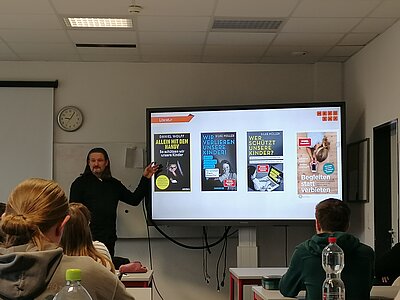 Eine Frau präsentiert verschiedene Plakate auf einem Bildschirm und davor sitzen mehrere Schüler in Sitzreihen.