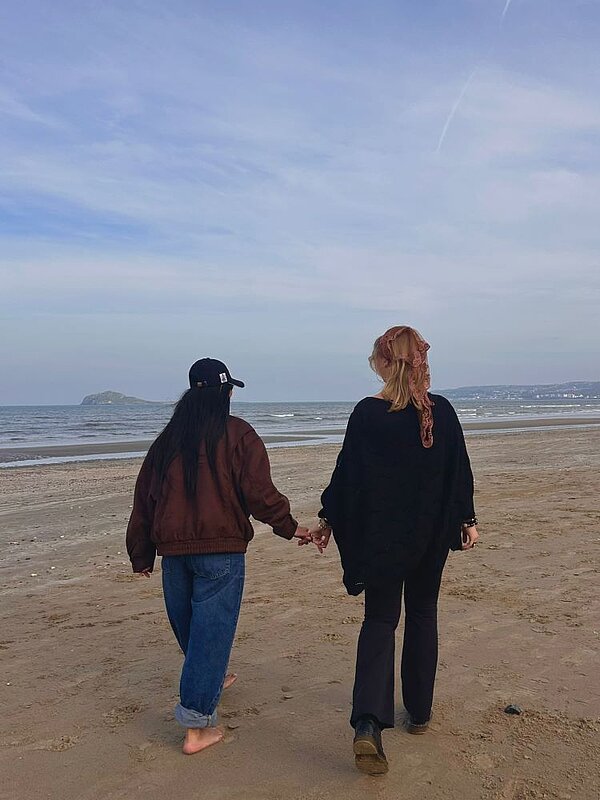 Zwei Personen sind von hinten zu sehen und gehen Hand in Hand am Strand entlang. Das Meer und der Horizont sind im Hintergrund sichtbar, während der Himmel leicht bewölkt ist. Die Stimmung wirkt entspannt und idyllisch.