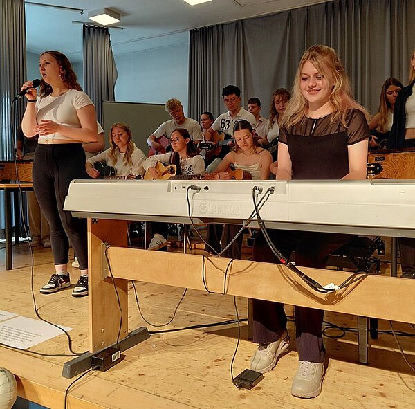 Herz. Humor. Haltung. Schulband auf der Bühne bei der Performance, im Vordergrund die Keyboarderin und eine Sängerin. Die Instrumentalisten musizieren stehend und sitzend im Hintergrund.