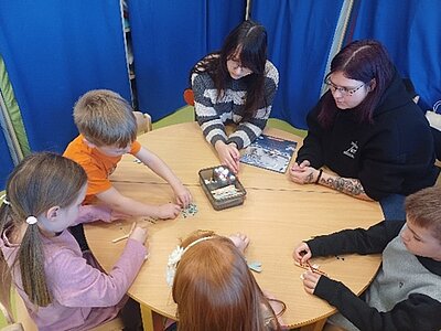 Mehrere Kinder und zwei erwachsene Auszubildende der Euro Akademie Dessau sitzen gemeinsam an einem runden Holztisch in einer Kita- oder Schulumgebung. Die Kinder basteln konzentriert mit bunten Perlen und Bastelmaterial, während die beiden angehenden Erzieherinnen unterstützend neben ihnen sitzen. Die Szene zeigt eine ruhige, kreative Aktivität im Rahmen des Bundesweiten Vorlesetags, bei dem die Auszubildenden Kindern Geschichten vorlasen und ergänzende pädagogische Angebote durchführten.