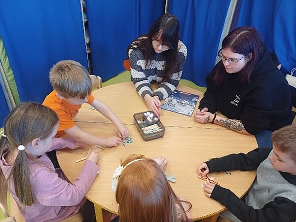 Mehrere Kinder und zwei erwachsene Auszubildende der Euro Akademie Dessau sitzen gemeinsam an einem runden Holztisch in einer Kita- oder Schulumgebung. Die Kinder basteln konzentriert mit bunten Perlen und Bastelmaterial, während die beiden angehenden Erzieherinnen unterstützend neben ihnen sitzen. Die Szene zeigt eine ruhige, kreative Aktivität im Rahmen des Bundesweiten Vorlesetags, bei dem die Auszubildenden Kindern Geschichten vorlasen und ergänzende pädagogische Angebote durchführten.
