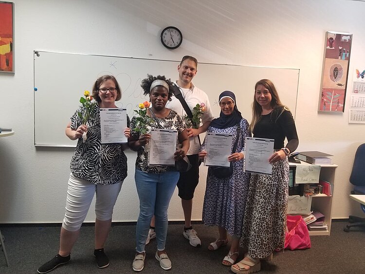Fünf Personen stehen nebeneinander im Klassenraum, halten stolz ihre Zertifikate in den Händen und lächeln. Drei der Personen halten zusätzlich eine Rose. Im Hintergrund ist eine weiße Tafel mit einer Uhr an der Wand zu sehen.