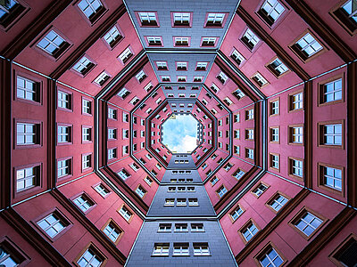 Blick nach oben in den achteckigen Innenhof des Palazzo Ottagonale in Berlin. Die rosaroten und grauen Fassaden mit gleichmäßig verteilten weißen Fenstern erzeugen eine markante, symmetrische Architektur. Der offene Himmel in der Mitte verstärkt die geometrische Tiefe des modernen Gebäudes.
