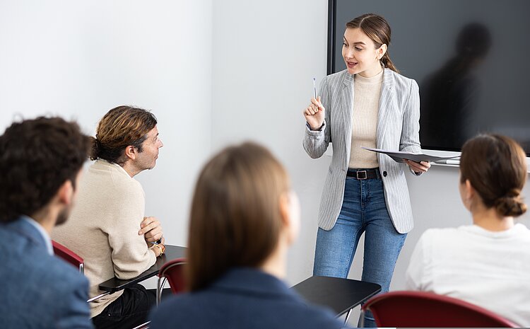 Berufspädagogische Pflichtfortbildung an der Euro Akademie Pößneck Zwei dunkelhaarige Frauen mit weißen Oberteilen und zwei schwarzhaarige Männer hören eine hell gekleideten Frau vor einer Tafel zu.