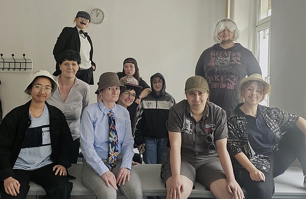 Die letzte Schulwoche der PFM 22: Ein unvergesslicher Abschluss Eine Gruppe von Personen sitzt und steht in einem Raum. Alle tragen verschiedene Hüte, Hemden und falsche Bärte oder Schnurrbärte – manche haben sich als Männer verkleidet. Im Hintergrund hängt eine Uhr an der Wand.