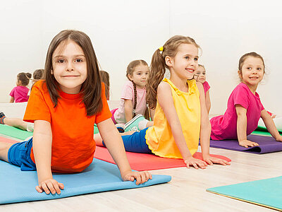 Das Bild zeigt Kinder auf bunten Matten in einem hellen Raum. Es handelt sich um eine Yoga- oder Sportklasse.