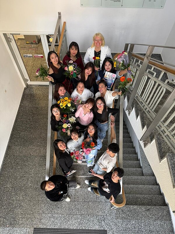Von oben aus der Vogelperspektive fotografierte Gruppe der Absolventinnen und Absolventen der Pflegefachkraft-Ausbildung, die auf einer Treppe im Gebäude der Euro Akademie Bitterfeld-Wolfen stehen. Viele tragen Blumensträuße und lächeln nach oben zur Kamera.