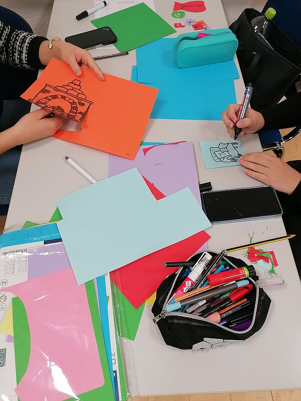 MeKoSax-Projekt „Trickfilmproduktion“ Auf einem Tisch liegen verschiedenfarbige Bögen Papier und Buntstifte. Mit Scheren Schneiden Schülerinnen Formen aus.