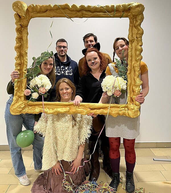 Lehrkraft und Studierende schauen lächelnd in ihren Faschingskostümen durch einen übergroßen selbst gestalteten Bilderrahmen und posieren für ein Foto.