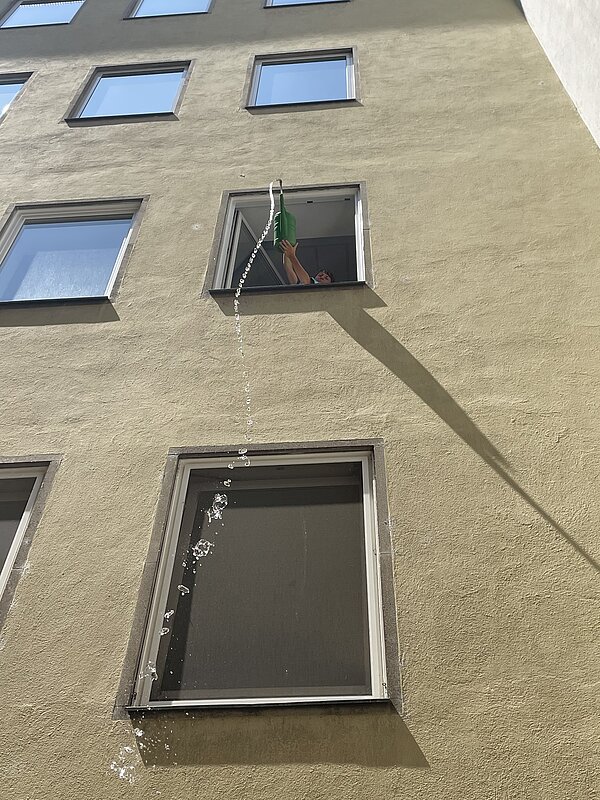 Eine Person gießt Wasser mit einer grünen Gießkanne aus einem Fenster in einem oberen Stockwerk eines mehrstöckigen Hauses. Der Wasserstrahl fällt in einem Bogen nach unten.