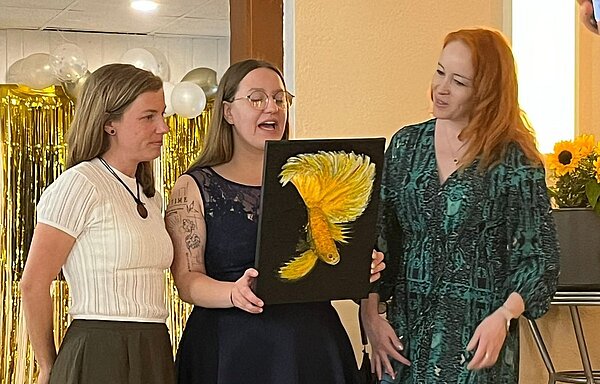 Feierliche Übergabe der Zeugnisse Zwei junge Mädchen, die links im Bild stehen, überreichen ein Bild an eine Lehrerin in einem grünen Kleid, die rechts steht. Auf dem Bild ist auf einem schwarzen Hintergrund ein gelber Fisch zu erkennen. Das Bild wird von dem Mädchen in der Mitte gehalten, das einen Text von der Rückseite des Bildes abzulesen scheint.