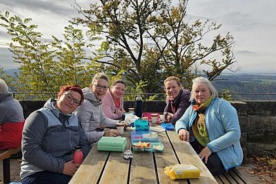 Eine Gruppe von Frauen sitzen in dicken Jacken draußen an einem Holztisch auf dem geöffnete Brotdosen mit Snacks liegen. Sie lächeln in die Kamera. Im Hintergrund sieht man einen Baum und eine weite bewaldete Landschaft.