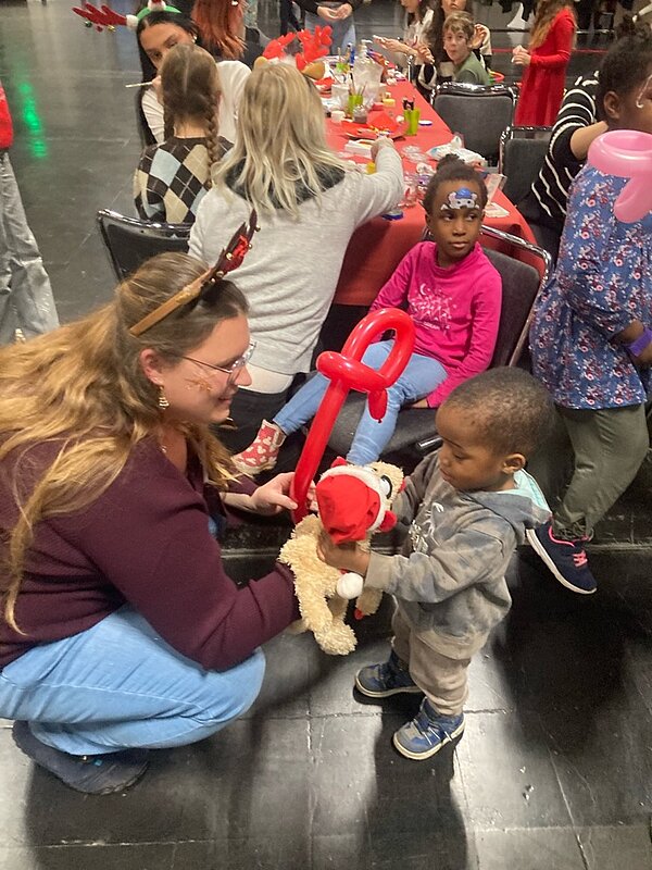 Eine Frau kniet neben einem kleinen Jungen und gibt ihm ein Stofftier mit einem roten Hut und einem roten Ballon in Form eines Schlüssels. Im Hintergrund sitzen mehrere Kinder an einem Tisch und scheinen an einer Veranstaltung teilzunehmen.