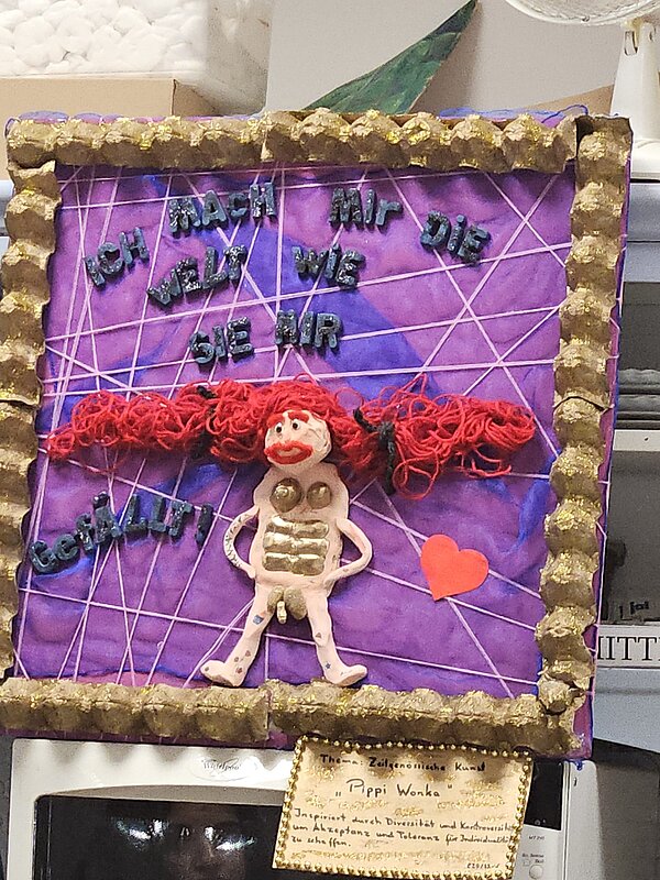 Ein Bilderrahmen mit rosa-lila Hintergrund und in dessen Mitte eine rothaarige Frau mit männlichen Geschlechtsteil abgebildet ist. Darüber steht "Ich mache mir die Welt, wie sie mir gefällt".