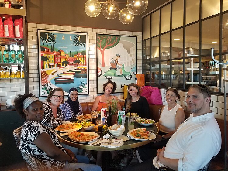 Eine Gruppe von sieben Personen sitzt fröhlich an einem Restauranttisch, auf dem große Pizzen, Salate und Getränke stehen. Im Hintergrund hängt bunte Italien-Dekoration an der Wand.