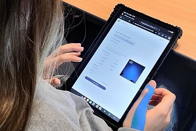 Eine Auszubildende mit langen, hellbraunen Haaren sitzt an einem Tisch und hält ein Tablet mit schwarzer Schutzhülle in beiden Händen. Auf dem Bildschirm ist eine Website geöffnet, die ein Formular mit mehreren Auswahlfeldern und einem kleinen Bild auf weißem Hintergrund zeigt. Die Person trägt einen grauen Pullover. Die Szene entstand im Rahmen eines Medienkompetenz-Projektes der Euro Akademie Görlitz zum Thema Fake News. Die Aufnahme vermittelt den Fokus auf digitale Mediennutzung und die kritische Auseinandersetzung mit Online-Inhalten.