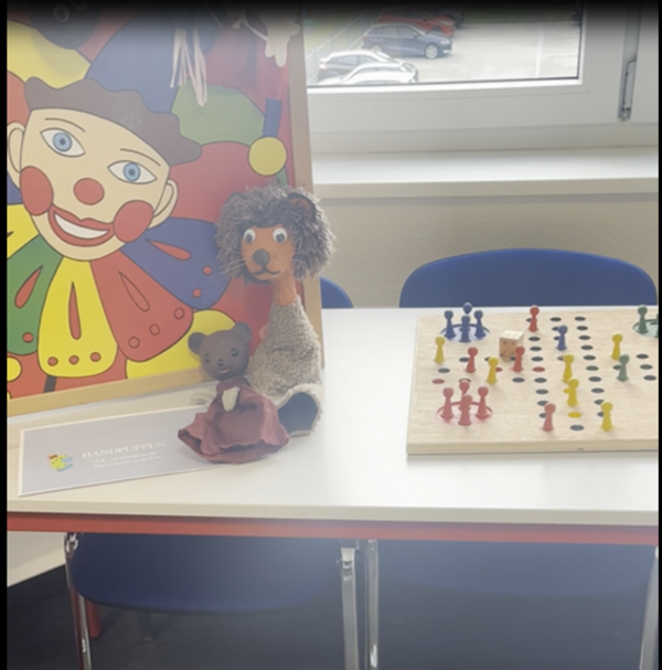 Auf einem weißen Tisch vor einem Fenster mit Blick auf einen Parkplatz stehen mehrere Gegenstände. Links ist ein buntes Bild eines Clowns mit roten Wangen, blauen Augen und einem mehrfarbigen Hut zu sehen. Vor dem Bild stehen zwei handgefertigte Stofftiere: eine kleine graue Maus mit rosa Kleid und eine größere Figur mit braunem Fell, schwarzer Mähne und langem Hals, die an ein Tier erinnert. Rechts auf dem Tisch liegt ein hölzernes Brettspiel mit bunten Spielfiguren in den Farben Rot, Gelb, Blau und Schwarz sowie einem kleinen Würfel. Im Hintergrund sind zwei blaue Stühle sichtbar. Die Szene wirkt wie eine Ausstellung oder Präsentation, passend zum Tag der offenen Tür an der Euro Akademie Zwickau.