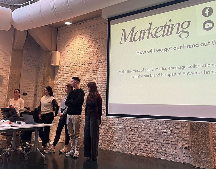 Eine Gruppe von fünf jungen Menschen steht vor einem großen Flatscreen und hält eine Präsentation über Marketingstrategien. Der Monitor zeigt den Titel „Marketing“ und Fragen zur Markenbekanntheit in englischer Sprache. Eine der Teilnehmenden spricht.