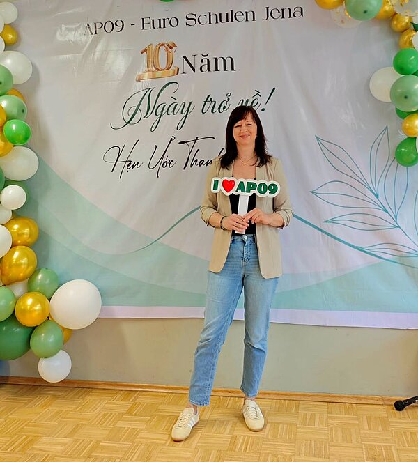 Eine Frau in beigem Blazer und blauer Jeans steht lächelnd vor einem Banner mit der Aufschrift „AP09 - Euro-Schulen Jena, 10 Năm, Ngày trở về!, Hẹn Uớc Thân”. Sie hält ein Schild mit der Aufschrift „I ❤️ AP09”. Links und rechts des Banners sind Luftballons in Grün, Gold und Weiß arrangiert.