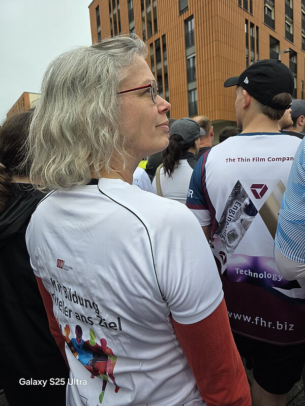 Nahaufnahme einer Frau mit grauen Haaren in Laufkleidung von hinten. Auf dem Shirt steht „Mit Bildung schneller ans Ziel“. Um sie herum sind weitere Läufer zu sehen.