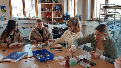 Vier weibliche Auszubildende sitzen an einem großen Tisch mit allerlei Bastel- und Maluntensilien, darunter Tuschkästen, Stifte und Karten.